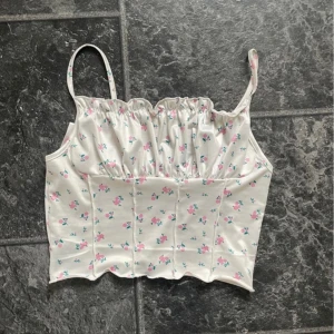 Vit blommig linnetopp - Säljer en superfin vit linnetopp med små rosa blommor och gröna blad. Toppen har smala axelband och en söt volangdetalj vid bysten. Perfekt för sommardagar eller en avslappnad fest. Den är i bra skick och väldigt bekväm att ha på sig!
