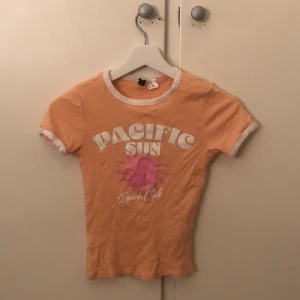 Orange t-shirt från H&M - Säljer en cool orange t-shirt från H&M med texten 'Pacific Sun Beach Club' i vitt och rosa. T-shirten har vita kanter vid ärmar och hals, och är kortärmad. Perfekt för en avslappnad stil!