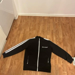Svart träningsset från Palm Angels - Snygg svart träningsjacka från Palm Angels med vita ränder längs ärmarna och dragkedja framtill. Jackan har en hög krage och Palm Angels-loggan på bröstet. Perfekt för en sportig look.