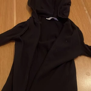 Svart hoodie med dragkedja - Säljer en svart hoodie med dragkedja och huva. Perfekt för en avslappnad stil. Den har långa ärmar och en enkel design som passar till det mesta.