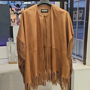 Brun läder poncho från Uterque - Snygg brun poncho i äkta läder från Uterque med fransdetaljer längs kanterna. Perfekt för att ge en bohemisk touch till din outfit. Ponchon har en öppen design utan knäppning. Lammskinn