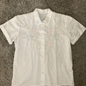 Vit blus/skjorta med broderade blommor - Vit loose fit kortärmad skjorta/blus med pastellfägad brodyr från 80-talet. Toppen är inköpt på Beyond retro och är i gott skick. Passar strl XS-M beroende på hur tight man vill att den sitter. Klicka gärna pả köp nu och meddela för eventuella frågor. :)