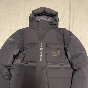 Svart dunjacka från Sail Racing - Snygg och funktionell svart dunjacka från Sail Racing med GORE-TEX teknologi. Jackan har en hög krage och justerbar huva för extra skydd mot väder och vind. Den är utrustad med flera praktiska fickor och dragkedja framtill. Perfekt för kalla dagar.