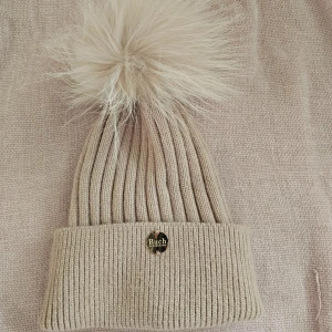 Beige ribbad mössa med pälsboll från Buch Copenhagen  - Snygg beige mössa från Buch  Copenhagen med avtagbar pälsboll i tvättbjörnsfuskpäls. Mössan är ribbad och tillverkad i 100% angora, vilket gör den både varm och mjuk. Perfekt för kyliga dagar!