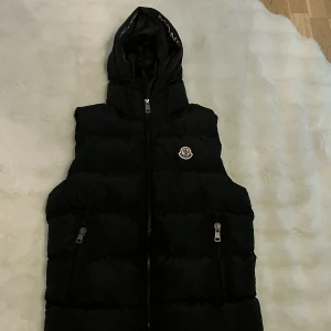Svart dunväst från Moncler - Snygg svart dunväst från Moncler med huva och dragkedja. Västen har två praktiska fickor med dragkedjor och Monclers logotyp på bröstet. Perfekt för kyligare dagar och för den kommande våren!