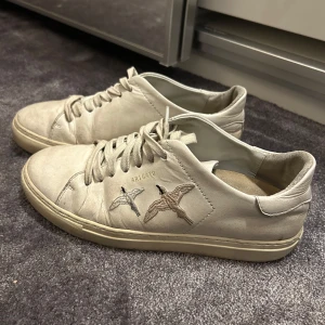 Beige sneakers från Axel Arigato - Snygga beige/ kräm vit sneakers från Axel Arigato med broderade fåglar på sidan. Skorna har snörning och är tillverkade i skinn, vilket ger en stilren och modern look. Tillkommer helt nya sulor!!
