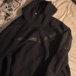 Svart hoodie från Nike - Säljer en svart hoodie från Nike med dragkedja och huva. Den har diskreta detaljer och en ficka på ärmen. Perfekt för en casual look.