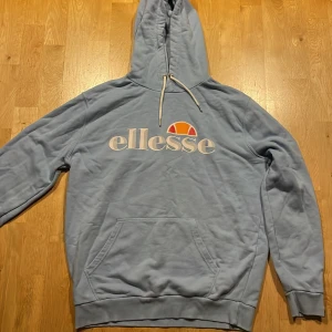 Ljusblå hoodie från Ellesse - Ljusblå hoodie från Ellesse med deras ikoniska logga i vitt och orange på bröstet. Tröjan har en stor ficka framtill och justerbar huva med snören. Pris kan diskutera