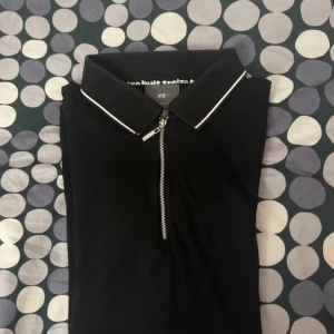 Svart pikétröja från H&M - Snygg svart pikétröja från H&M i slim fit med dragkedja framtill. Tröjan har korta ärmar och vita detaljer på krage och ärmslut. Perfekt för en stilren look.