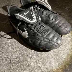 Svarta fotbollsskor från Nike - Säljer ett par svarta Nike fotbollsskor med snörning och gulddetaljer på sulan. Skorna har en quiltad design och är perfekta för fotbollsplanen.