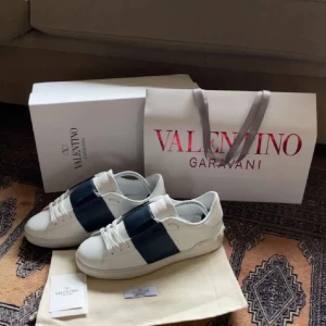 Valentino Garavani sneakers i vitt och blått - Snygga Valentino Garavani sneakers i vitt med en bred blå rem över ovansidan. Skorna har en klassisk design med snörning och en bekväm sula. Perfekta för en stilren look.