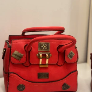 Röd handväska med detaljer - Snygg röd handväska med guldfärgade detaljer och dekorativa märken. Väskan har ett handtag och en stilren design som passar perfekt för att ge en färgklick till din outfit.