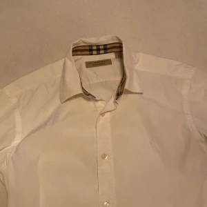 Vit skjorta från Burberry - Säljer en stilren vit skjorta från Burberry med klassiskt rutigt mönster på insidan av kragen och ärmsluten. Skjortan har långa ärmar och knappar. Priset är inte hugget i Sten