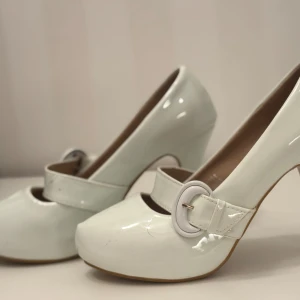 Vita pumps med spänne - Eleganta vita pumps med rundad tå och dekorativt spänne över foten. Skorna har en hög klack och är perfekta för en stilren look.
