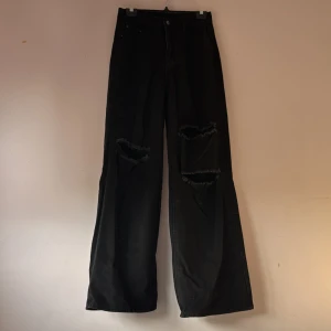 Svarta byxor med slitningar - Snygga svarta byxor med slitningar på knäna. De har en loose passform och är perfekta för en avslappnad stil. Byxorna är högmidjade och har en klassisk design med dragkedja och knapp.