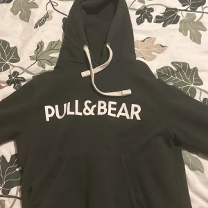 Grön hoodie från Pull&Bear - Säljer en grön hoodie från Pull&Bear med vit text på framsidan. Den har en klassisk känguruficka och justerbar huva med vita snören. Perfekt för en avslappnad stil.