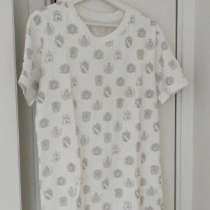 Thomas Burberry t-shirt - Snygg vit t-shirt från Thomas Burberry med ett unikt mönster i grått. Tillverkad i 100% bomull för en bekväm känsla. Perfekt för en stilren och avslappnad look. Knappt använd, max en gång.