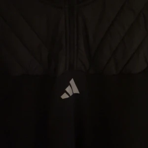 Svart jacka från Adidas - Snygg svart jacka från Adidas med quiltad design upptill och klassiska vita ränder längs ärmarna. Jackan har en dragkedja framtill och är perfekt för en sportig look.