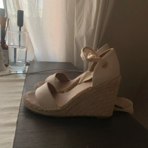 Beige kilklack med knytband - Snygga beige sandaler med kilklack och knytband runt ankeln. Perfekta för sommarens alla tillfällen. Klacken är flätad och ger en bohemisk känsla. Använd endast en gång.