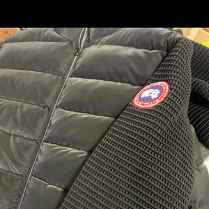 Svart dunjacka med stickade ärmar - Snygg svart dunjacka med stickade ärmar och ett runt märke på ärmen. Jackan har en dragkedja framtill och quiltad design som ger en modern look. Perfekt för kyligare dagar. det finns många storlek av denna jag kan priset kan diskuteras