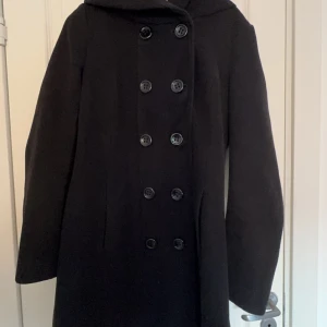 Svart kappa med knappar - Elegant svart kappa med dubbelknäppning och långa ärmar. Perfekt för kyligare dagar med sin stilrena design och praktiska knappar framtill.
