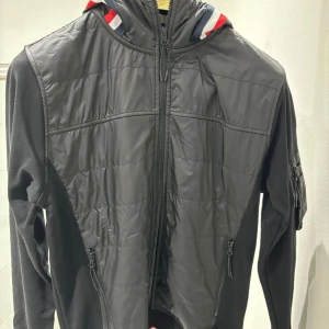 Svart jacka med huva från Moncler - Snygg svart jacka från Moncler med huva och dragkedja. Jackan har långa ärmar och ett broderat Moncler-märke på ärmen. Den är tillverkad i bomull och har detaljer i rött, vitt och blått på huvan.