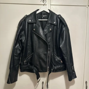 Svart skinnjacka från Missguided - Snygg svart skinnjacka från Missguided med dragkedja och bälte i midjan. Jackan har långa ärmar och axelklaffar för en cool look. Perfekt för att ge din outfit en edgy touch.