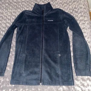 Svart fleecejacka från Columbia - Säljer en svart fleecejacka från Columbia med dragkedja framtill. Jackan har en hög krage och långa ärmar, perfekt för kyliga dagar. Tillverkad i 100% polyester för extra värme och komfort.             Köpt på Humana(Mariatorget) där ordinarie pris var 500kr.