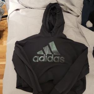 Svart hoodie från Adidas - Säljer en svart hoodie från Adidas med det klassiska logotyptrycket i silver på framsidan. Hoodien har en bekväm passform med långa ärmar och en praktisk huva. Perfekt för en avslappnad stil.Den är en magtröja men bra skick på tröjan lappen ser man knappt vad det står på. Köparen står för frakt