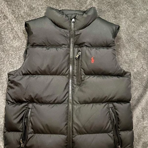 Svart dunväst från Polo Ralph Lauren - NYSKICK! Köptes oktober 2024. Knappt använd. Säljer en snygg svart dunväst från Polo Ralph Lauren. Västen har en dragkedja framtill och två praktiska fickor med dragkedjor. Den är perfekt för kyligare dagar och har en liten röd logga på bröstet.