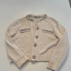Beige kofta från Zara med pärldetaljer - Säljer en elegant beige kofta från Zara med pärldetaljer vid halsringning och fickor. Koftan har långa ärmar och knäpps med dekorativa knappar. Perfekt för att ge en touch av glamour till din outfit.