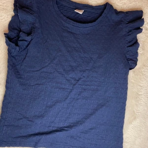 Blå topp från Lindex - Säljer en söt mörkblå topp från Lindex med volangdetaljer vid ärmarna. Toppen har en fin struktur i tyget och är perfekt för en avslappnad stil. Passar bra till både jeans och kjol. Perfekt för sommarens äventyr! 💙 OBS ljuset gör att den ser lite blåare ut! Liten i storlek 