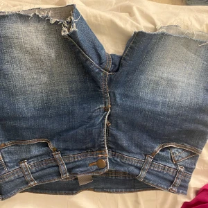 Blå jeansshorts med slitna detaljer - Säljer ett par coola blå jeansshorts från Wrangler med slitna detaljer och klassiska femficksdesign. Perfekta för en avslappnad stil. De har en knapp och dragkedja framtill.