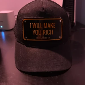 Svart keps från John Hatter & Co - Snygg svart keps med texten 'I WILL MAKE YOU RICH' på framsidan. Märket John Hatter & Co är broderat på insidan. Perfekt för en cool och självsäker stil.