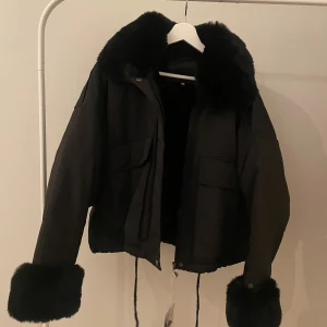 Svart jacka med pälsdetaljer - Snygg svart jacka med fluffiga pälsdetaljer på krage och ärmslut. Jackan har en dragkedja framtill och två stora fickor. Perfekt för kyliga dagar när du vill hålla stilen.