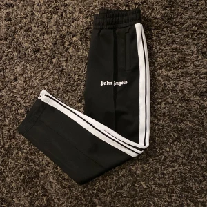 Svarta byxor från Palm Angels - Snygga svarta byxor från Palm Angels med vita ränder längs sidorna. Byxorna har en elastisk midja och sidofickor. Perfekta för en avslappnad stil med en touch av streetwear.