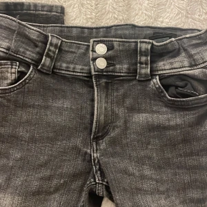 Grå bootcut jeans från H&M - Snygga grå bootcut jeans från H&M med justerbar midja och låg midja. De har dubbla knappar fram och fickor med lock och knapp bak. Perfekta för en avslappnad stil. Frakt 40kr