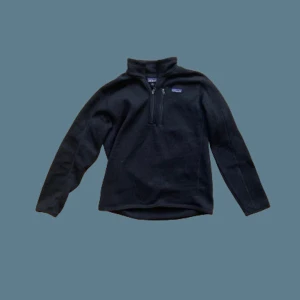 Svart fleece tröja från Patagonia - Säljer en svart fleece tröja från Patagonia med halv dragkedja. Perfekt för kyliga dagar med sin mjuka och varma känsla. Tröjan har långa ärmar och en hög krage för extra värme. Passar bra till både vardag och friluftsliv.