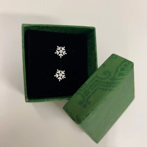 Snöflingeformade örhängen i äkta silver  - Eleganta örhängen i form av snöflingor, tillverkade i silver. Perfekta för att ge en gnistrande touch till din outfit. Kommer i en vacker grön ask med mönster. Äkta silver 925