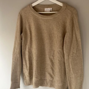 Beige stickad tröja från Vila - Säljer en mysig beige stickad tröja från Vila. Tröjan har långa ärmar och ribbstickade muddar vid ärmslut och nederkant. Perfekt för kyliga dagar!