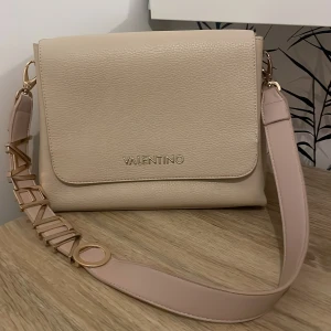 Beige axelväska från Valentino - Snygg beige axelväska från Valentino med justerbar axelrem och gulddetaljer. Väskan har en stilren design med en praktisk dragkedja inuti för säker förvaring. Perfekt för att bära dina dagliga nödvändigheter med stil.