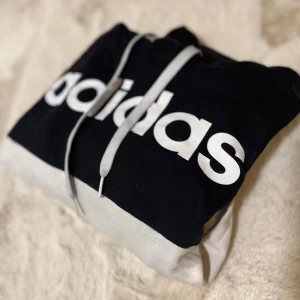 Svart och vit hoodie från Adidas - Säljer en snygg svart och vit hoodie från Adidas med stor logga på framsidan. Tröjan har långa ärmar med vita ränder längs sidorna och en praktisk huva med dragsko. Perfekt för en sportig look!