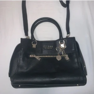 Svart handväska från Guess - Snygg svart handväska från Guess i skinn med silverfärgade detaljer och dragkedja. Väskan har både handtag och en justerbar axelrem. Perfekt för att bära dina dagliga nödvändigheter med stil. Inuti finns flera fack för enkel organisering.