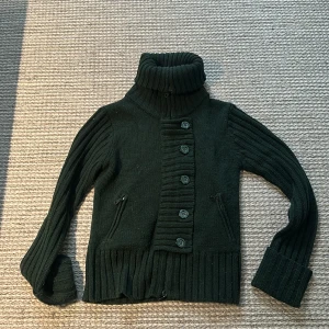 Mörkgrön stickad kofta med polokrage - Säljer en mysig mörkgrön stickad kofta med polokrage och knappar framtill. Koftan har långa ärmar och ribbade detaljer vid ärmslut och nederkant. Perfekt för kyliga dagar! Inga defekter eller noppror, köpt på humana