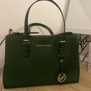 Grön handväska från Michael Kors - Snygg grön handväska från Michael Kors  med gulddetaljer. Väskan har dubbla handtag och en avtagbar axelrem. Den stängs med dragkedja och har ett rymligt inre med flera fack. Perfekt för att bära dina dagliga nödvändigheter med stil.