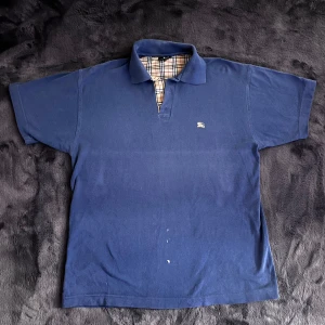 Burberry Pike - Säljer en klassisk blå pikétröja från Burberry med korta ärmar och en stilren krage. Insidan av kragen har det ikoniska Burberry-rutiga mönstret. Perfekt för en avslappnad men stilfull look. Skick 7/10, fläck på magen som säkert går veck med nått medel. Storlek XL men passar L. Pris går att diskutera ✅