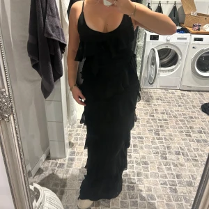 Balklänning med volanger  - Elegant svart långklänning med volanger och smala axelband. Perfekt som balklänning med en slits vid ena benet. Helt ny fortfarande med prislapp.