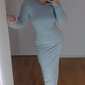 Ljusgrå långklänning med öppen rygg - Snygg ljusgrå långklänning med en elegant öppen rygg. Klänningen har en tight passform och långa ärmar, perfekt för en stilren look. Materialet ser mjukt och bekvämt ut, vilket gör den idealisk för både vardag och speciella tillfällen.