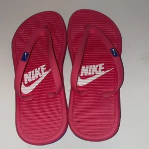 Rosa flip-flops från Nike - Snygga rosa flip-flops från Nike med vit logga på sulan. De har en blå sula med rutmönster undertill för bra grepp. Perfekta för sommardagar vid stranden eller poolen.