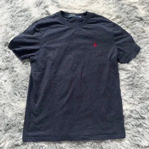 Mörkblå t-shirt från Ralph Lauren - Säljer en stilren mörkblå t-shirt från Ralph Lauren med det klassiska röda broderade logotypen på bröstet. Perfekt för en avslappnad och snygg look. T-shirten är kortärmad och gjord i mjukt bomullsmaterial.